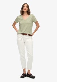 Grøn t-shirt med V-hals og korte ærmer, parret med hvide jeans og et flettet brunt bælte. Modellen har mørke loafers på; neutral baggrund.