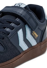 Gros plan sur une basket bleu marine avec une bride auto-agrippante, des rayures chevron bleu clair, des lacets noirs et une semelle marron, présentant le logo "hummel" et le texte "HANDBALL PERFECT".