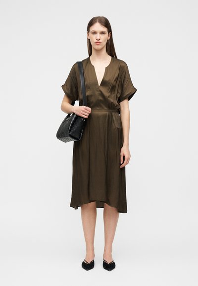 Γυναίκα με καφέ φόρεμα μέχρι το γόνατο, μαύρα μυτερά παπούτσια και μαύρη τσάντα ώμου με υφή, σε απλό φόντο.