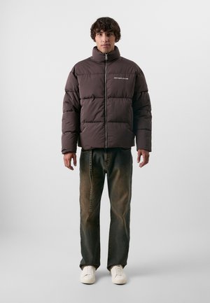 Καφέ φουσκωτό jacket με καπιτονέ σχέδιο και ψηλό γιακά. Διαθέτει μπροστινό φερμουάρ και κείμενο "ΜΗ ΣΤΕΡΕΙΣ ΤΗΝ ΚΟΥΛΤΟΥΡΑ". Συνδυάζεται με σκούρα τζιν και άσπρα αθλητικά παπούτσια.