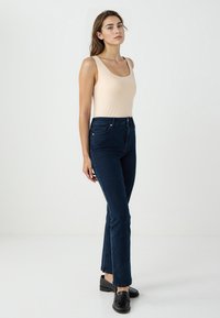 Donna che indossa un top beige senza maniche, jeans a vita alta blu scuro e mocassini neri, in piedi contro uno sfondo chiaro e semplice.