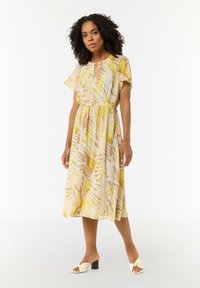 Mehrfarbiges langes Kleid in Gelb und Beige mit einem Blättermuster, kurzen schwingenden Ärmeln und einer taillierten Taille. Kombiniert mit cremefarbenen Absatzsandalen.