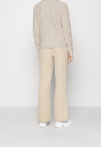 Personne debout de dos, portant un pull côtelé beige, un pantalon large beige à rayures verticales blanches, et des baskets blanches sur un fond uni.