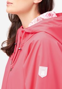 Vattentät rosa regnjacka med mönstrat huva foder, silverfärgad hårdvara och en vit logotypapplikation på ärmen.