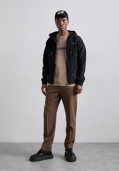 Sudadera negra con cremallera y cordones, camiseta beige con texto, pantalones marrones ajustados y zapatillas de plataforma negras. Gorro negro con logo.