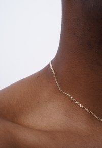Close-up of dark skin neck and shoulder wearing a thin gold chain necklace against a plain white background.