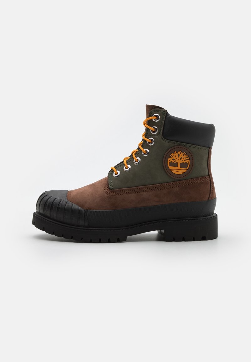 Botte à lacets marron et vert olive avec col rembourré noir, semelle robuste, embout noir et patch logo orange sur le côté.