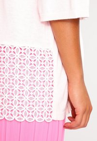 Bílý pletený top s texturovanou růžovou krajkovou částí na spodním lemu. Krajka má oválný vzor. Krátké rukávy s žebrovaným zakončením.