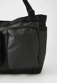Bolsa tote negra con un acabado mate y suave, y correa de hombro ajustable. Presenta dos bolsillos frontales y costuras duraderas a lo largo de las costuras.