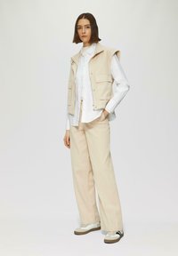 Gilet sans manches beige avec poches avant, porté sur une chemise blanche à boutons et un pantalon large crème. Des baskets blanches et marron complètent le look.