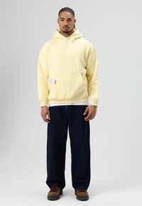 Geel hooded sweatshirt met een voorzak, gecombineerd met donkerdenim broek en bruine veterlaarzen, ontworpen voor een ontspannen pasvorm.