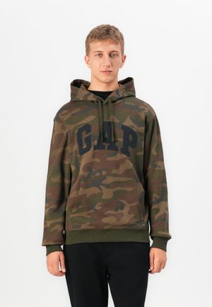 Un jeune homme portant un sweat à capuche camouflage vert et marron avec un logo GAP noir et un pantalon noir, se tenant devant un fond blanc uni.