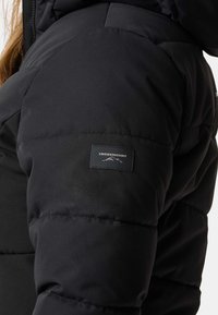 Veste isolée noire avec un design matelassé, tissu texturé et une étiquette de marque sur la manche avec un graphisme de montagne.