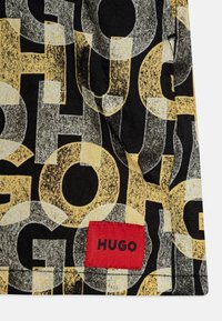 Trumpetinis marškinėliai su juoda medžiaga, puikuojančia geltonais ir pilkais abstrakčiais rašmenimis. Raudonas etiketė su "HUGO" akcentu arti apačios.