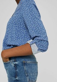 Camicia azzurra con pois bianchi e maniche arrotolate; il tessuto sembra morbido. Indossata con jeans in denim, evidenziando un fit casual.