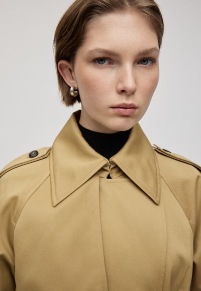 Trench coat beige con ampio colletto, dettagli con bottoni e spalline. Tessuto testurizzato e dolcevita nera visibile sotto.