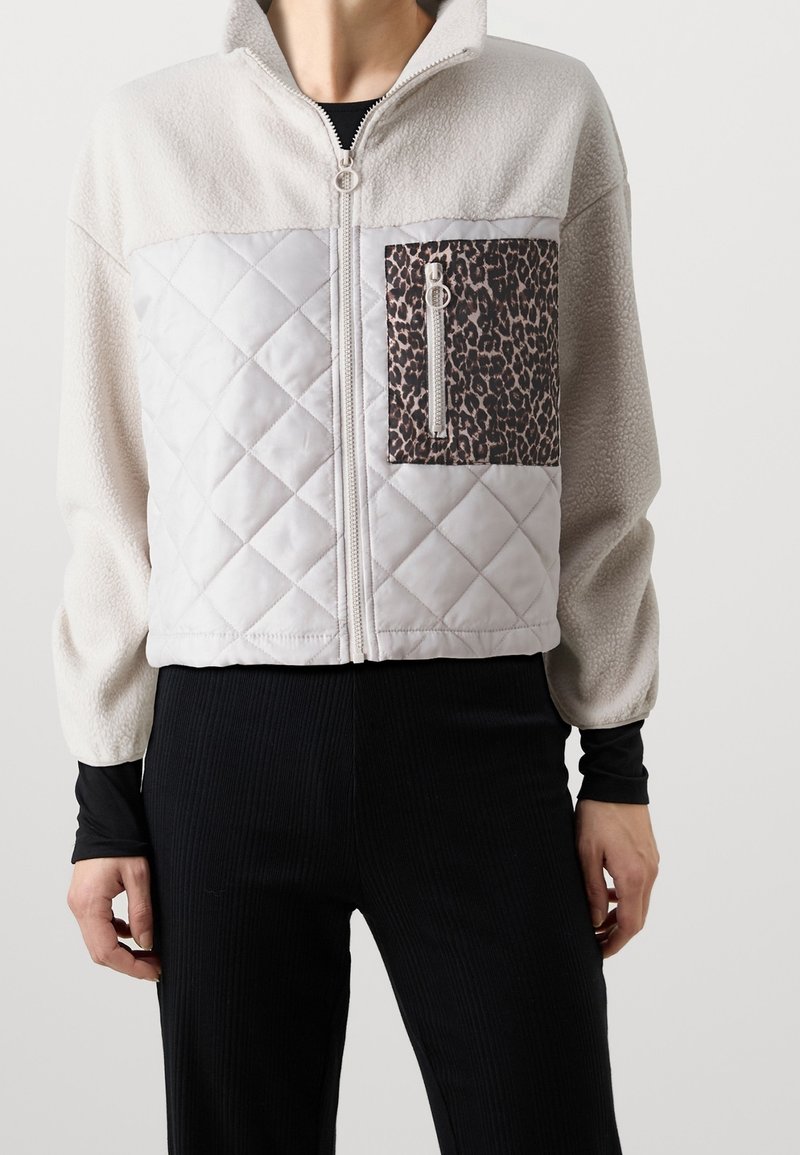 Vaaleanharmaa vetoketjullinen takki, jossa on tikattu etuosa ja leopardia kuvioiva paikkatasku. Valmistettu pehmeästä fleecemateriaalista.