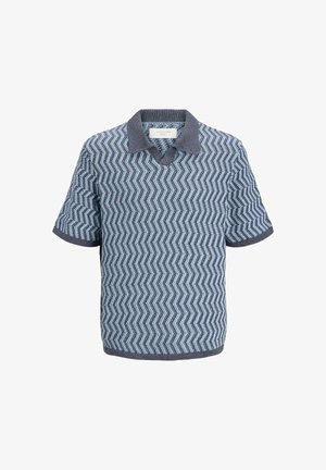 Adolescent aux cheveux bouclés portant une chemise bleue à manches courtes à motif zigzag, un pantalon noir à jambes larges et des baskets blanches, debout et souriant.