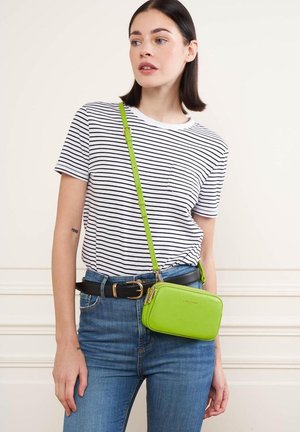 Sac en cuir vert à bandoulière avec fermeture éclair, sangle réglable et texture lisse. Le modèle porte une chemise rayée et un jean en denim.