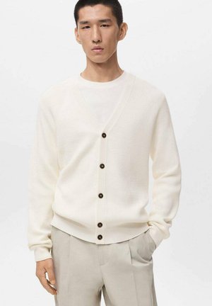 White knit cardigan with a V-neck, brown buttons, ribbed texture, and long sleeves, paired with a white underlayer and beige trousers.