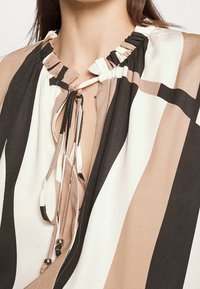 Halenka s kulatým, volánkovým výstřihem, zdobená svislými pruhy v odstínech béžové, hnědé a krémové. Zapínání zvýrazněné ozdobnými korálky.