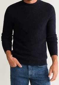 Uomo che indossa un maglione lungo a maniche nere con texture e jeans blu, con una mano in tasca e l'altra rilassata al suo fianco.