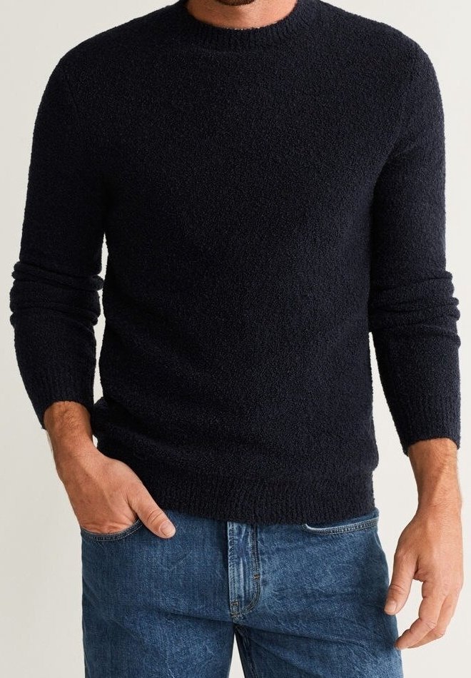 Uomo che indossa un maglione lungo a maniche nere con texture e jeans blu, con una mano in tasca e l'altra rilassata al suo fianco.