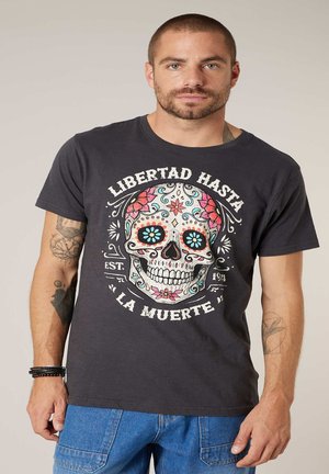 Homme portant un t-shirt noir avec un crâne sucré coloré et le texte espagnol « Libertad Hasta La Muerte » ainsi qu'un jean bleu, debout devant un fond uni.