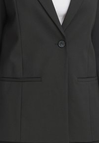 Veste noire à un bouton avec revers crantés et deux poches frontales horizontales, portée sur un haut blanc.