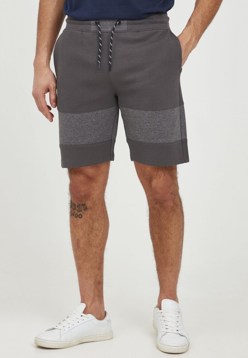 Grå sweatshorts med et teksturert grått panel, snor i livet og sidelommer, kombinert med hvite joggesko.