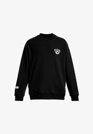 Sudadera negra con cuello redondo y mangas largas, con una pequeña cara de oso de peluche gris bordada en el pecho izquierdo y una etiqueta en el puño de la manga izquierda.
