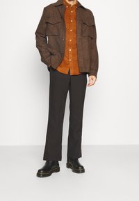 Chaqueta a cuadros marrón sobre una camisa naranja, combinada con pantalones negros y botas negras resistentes. Incluye bolsillos frontales y un diseño de botones.