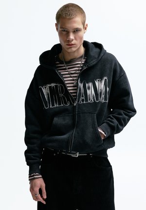 Fekete cipzáras kapucnis pulóver, melyen fehérrel a "NIRVANA" felirat látható a mellrészen. Alatta csíkos hosszú ujjú ing, fekete kordbársony nadrággal párosítva.