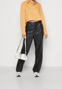 Pantalon large en simili cuir noir, associé à un haut orange court. Un petit sac à main blanc au design épuré et aux accents noirs est tenu.