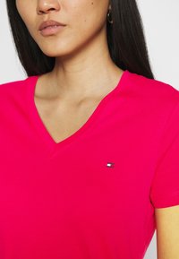 Camiseta fucsia de cuello en V hecha de algodón, con mangas cortas y un pequeño logo bordado en el pecho. Textura suave.
