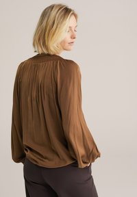 Blond kvinna med stängda ögon iförd en brun blus med lång ärm och veck samt mörka byxor, stående mot en beige bakgrund.