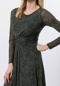 Schwarzes und grünes gemustertes Kleid mit langen, durchsichtigen Ärmeln, gerafftem Taillenbereich und fließendem Rock. Texturiertes Gewebe mit dezentem Schimmer.