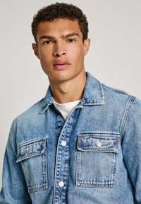 Chaqueta de denim en azul claro con dos bolsillos en el pecho, herrajes plateados, cierres de botón y un cuello clásico. Tejido texturizado con ligero desgaste.