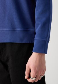 Sudadera azul marino con puños y dobladillo acanalados. El material muestra una leve textura; combinada con pantalones negros. Mano adornada con anillos de plata.