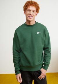 Sudadera verde de Nike con cuello redondo, puños y dobladillo acanalados. Presenta un pequeño logo blanco en el lado izquierdo del pecho. Llevada con jeans oscuros.