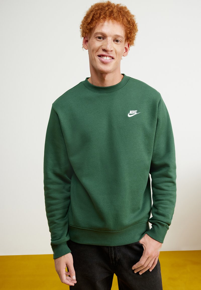Sudadera verde de Nike con cuello redondo, puños y dobladillo acanalados. Presenta un pequeño logo blanco en el lado izquierdo del pecho. Llevada con jeans oscuros.
