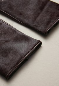Dunkelbrauner Kunstpelzstoff mit einer glatten Textur, abgerundeten Kanten und kontrastierenden Lederrändern entlang der Nähte.