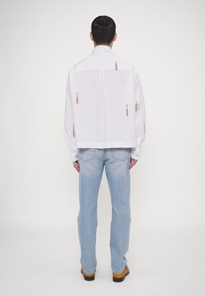 Hvid skjorte med knaplukning og kort design, der har flere orange lynlåsdetaljer på ryggen. Båret med lyseblå jeans og brune sko.