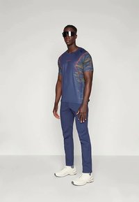 Jūras spēku sporta krekls ar rakstainām piedurknēm, sarkaniem akcentiem, kopā ar atbilstošām jūras spēku biksēm un baltām kedām. Modelī ir tumšas saulesbrilles.