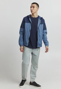 Giacca a vento blu navy e azzurro chiaro con cappuccio regolabile, polsini elasticizzati e zip completa. Abbinata a una maglietta blu navy e jeans azzurro chiaro.