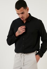 Camisa negra de botones con una tela texturizada, de mangas largas y con cuello. Presenta botones oscuros y se combina con pantalones de color claro.
