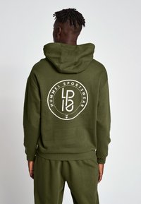 Oliwkowa bluza z kapturem z dużym białym okrągłym logo na plecach, zawierającym tekst i geometryczne wzory. Wykonana z miękkiego materiału.