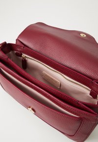 Red textured leather handbag open to show beige interior, zippered compartment, and brand label "Coccinelle Authentic Italian Leather."
