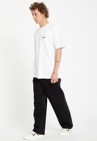 White cotton t-shirt with short sleeves and black embroidery on the chest, paired with black relaxed-fit pants and white sneakers.