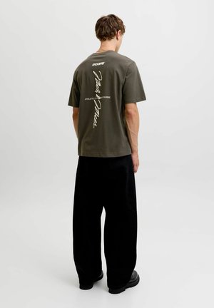 Jeune homme debout, dos à la caméra, portant un t-shirt vert olive foncé avec un texte vertical blanc, un pantalon noir ample et des chaussures noires.